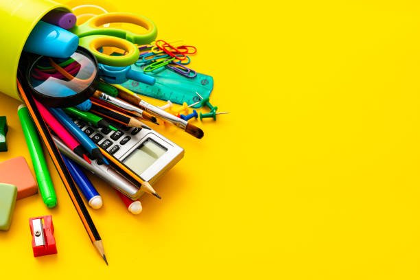 Close up view of a desk organizer full of school supplies laying on yellow background. Copy space. The composition includes a drawing compass, color pencils, paint brushes, paper clips, pencil sharpener, eraser, crayons, pocket calculator, thumbtack, rubber bands, notepad, stapler, adhesive tape, felt tip pen and scissors.